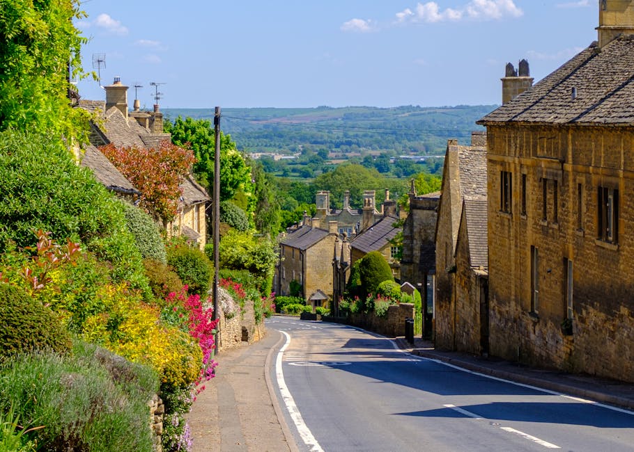 A scenic view of a quiet, gently curving rural street in Cowley, featuring stone cottages with slate roofs on either side. The gardens bordering the road are lush and vibrant, with a variety of flower