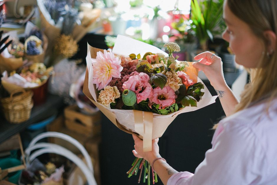 A woman holding a bouquet arrangement of fresh flowers, including large pink dahlias, pink roses, light beige carnations, and dark green foliage, wrapped in cream and white paper with a satin ribbon.