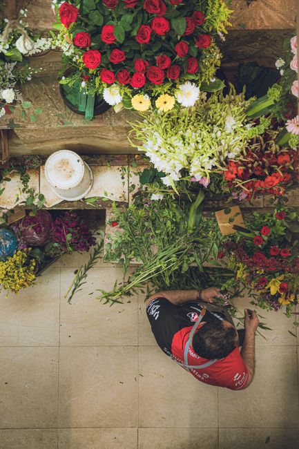 An overhead view of a florist arranging a floral gift display featuring a bouquet of red roses with green foliage, complemented by white daisies and yellow chrysanthemums. The arrangement is placed on